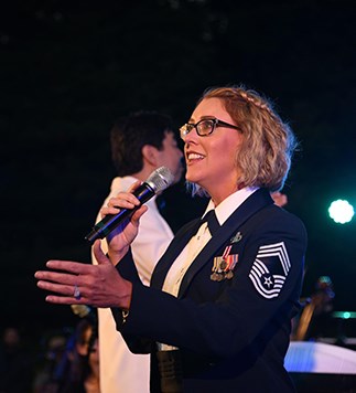 CMSgt Christine Germain A woman with short curly blonde hair, and glasses, wearing a blue United States Air Force Dress uniform sings at an outdoor amphitheater at night.