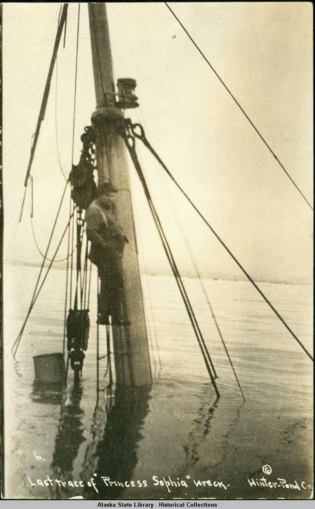 Man standing on foremast of a sunken ship in water.