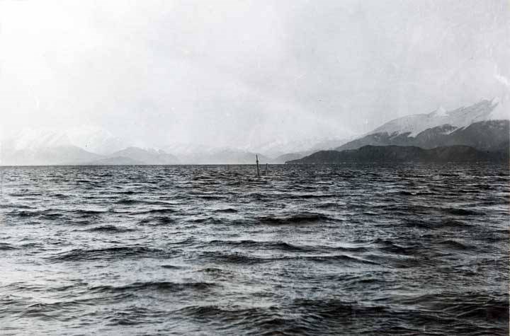 Image of sunken ship with tip of ship mast sticking out of a large body of water. Mountains in the background.
