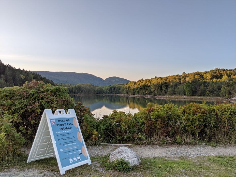 a sign saying help us study fall foliage on a path in front of pond with mountains in the distance