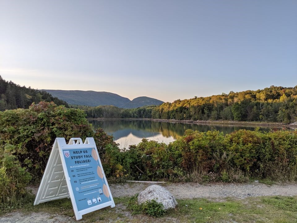 Photo Station a sign saying help us study fall foliage on a path in front of pond with mountains in the distance