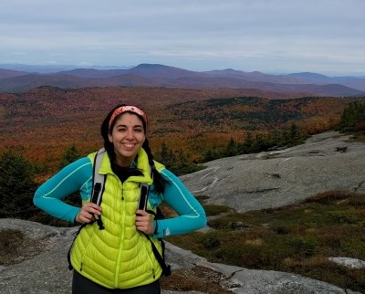 female in blue shirt and yellow vest on a mountain summit with fall colors visible on trees