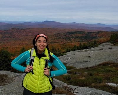 researcher profile female in blue shirt and yellow vest on a mountain summit with fall colors visible on trees