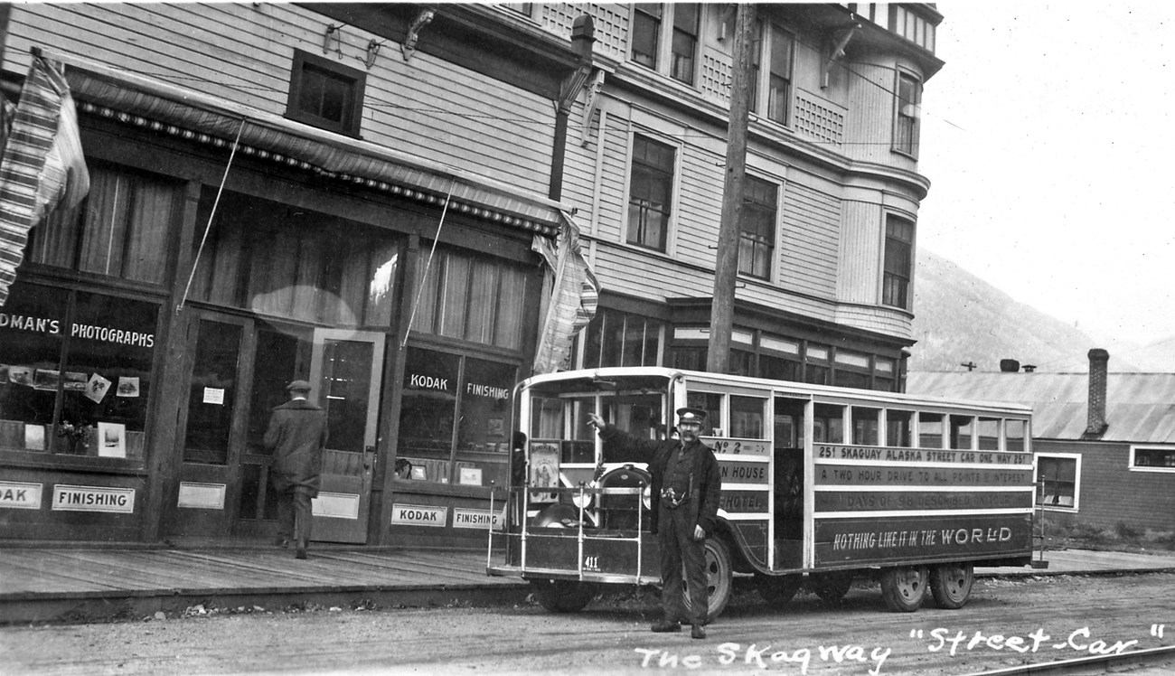Man in uniform in front of streetcar.  He is pointing.  A large building is in background.