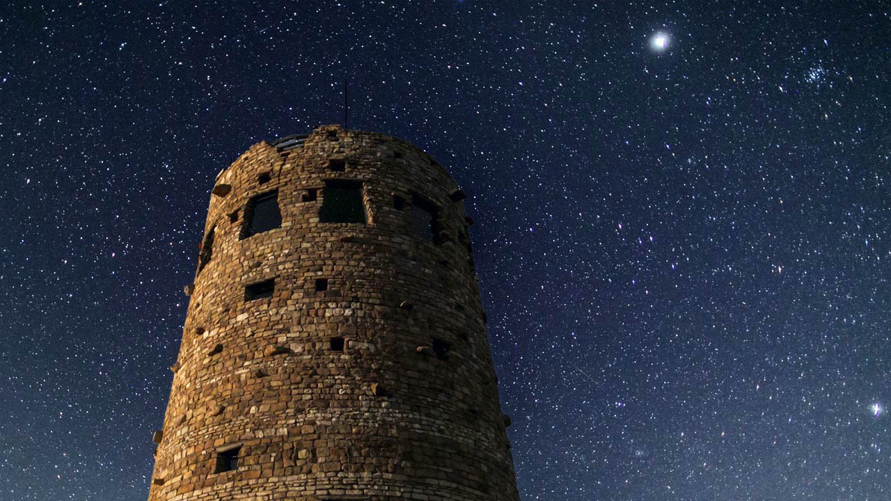 Perched on the rim of Grand Canyon, the Watchtower reflects the architecture of the ancestral Puebloan Indians of the Four Corners region. Climb the stairs for views up and down the canyon. . On a clear day, one can see well over 100 miles. (161 km) Stars in a night sky above a circular stone tower with windows of different sizes around the circumference.