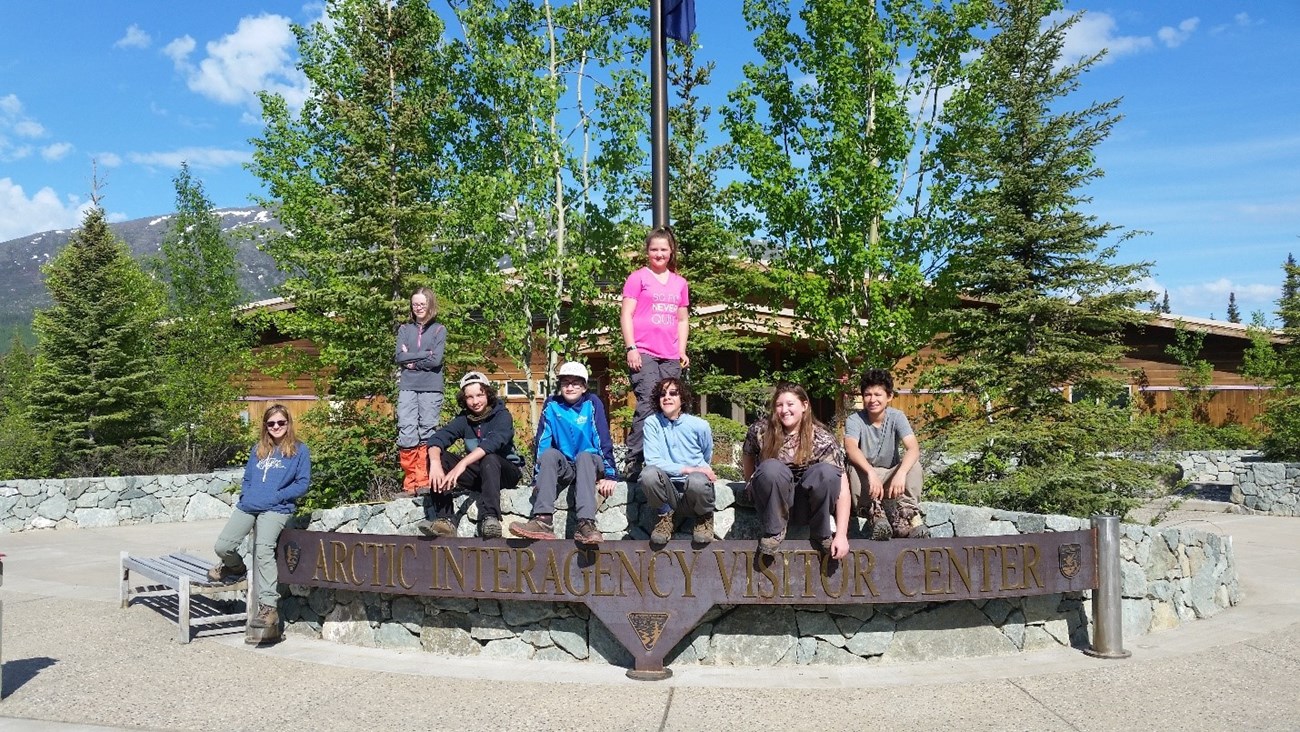 A group of eight students sit atop the Arctic Interagency Visitor welcome sign on a sunny day.