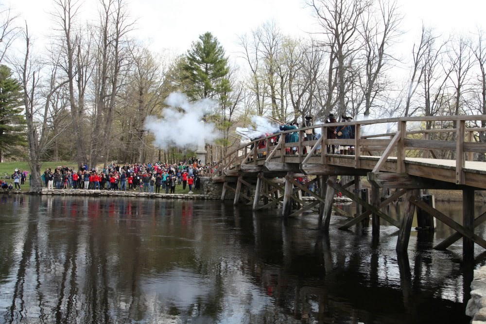 A wooden bridge stretches across the water while reenactors fire guns.