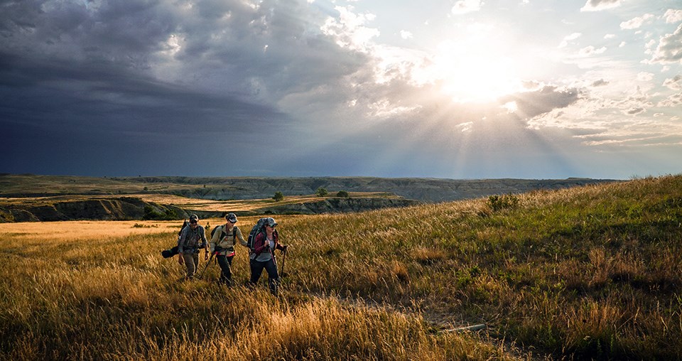 Three people hiking across a brown grass prairie with badlands in the background
