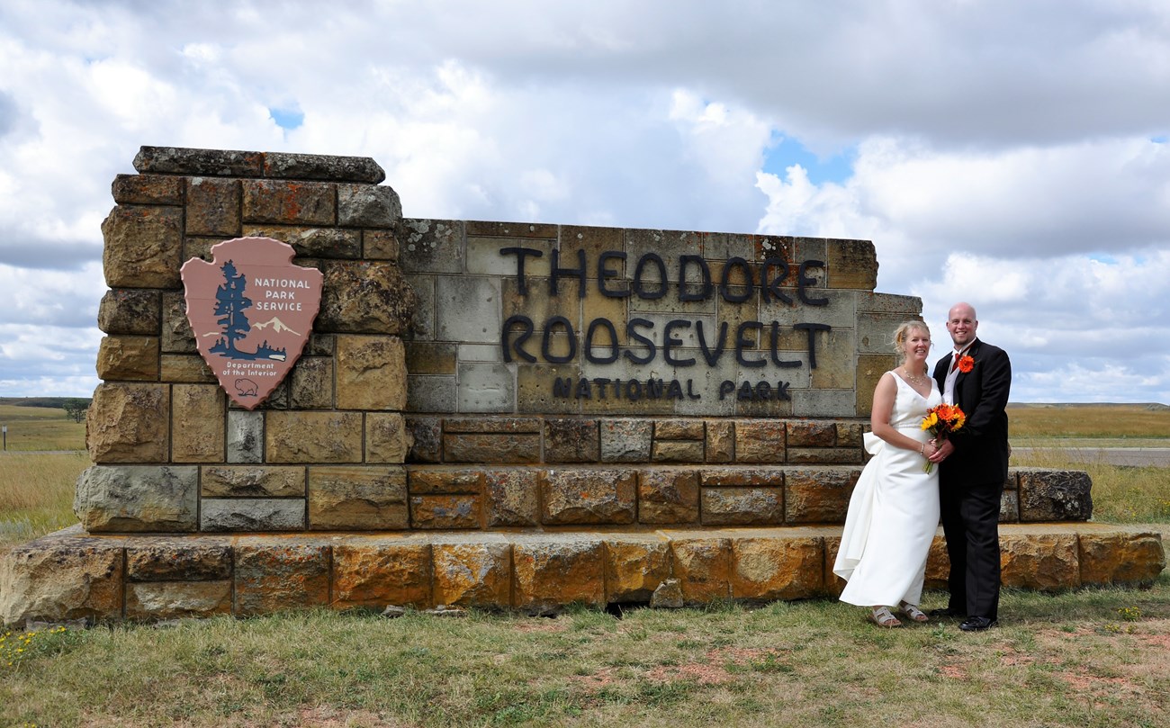 a bride and groom stand in front of the Thedore Roosevelt National Park Sign.
