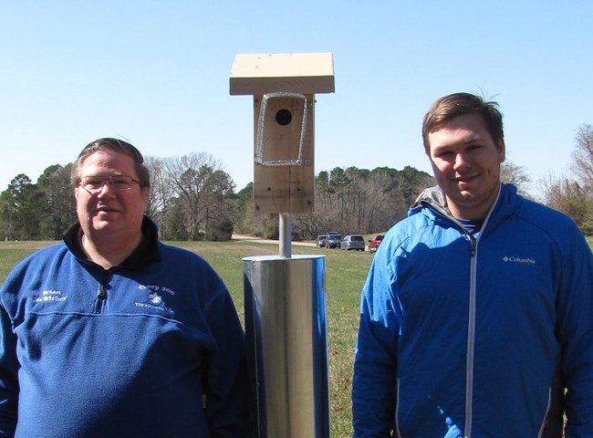 Two men in blue jackets stand next to a bluebird house.