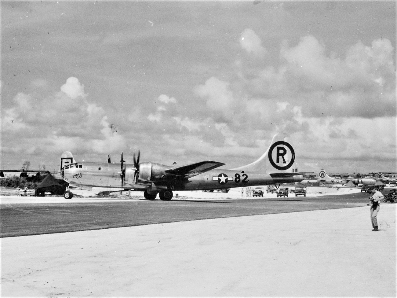 Enola Gay bomber after strike at Hiroshima, entering Tinian airfield Large metallic plane with propellers on sandy runway, various military vehicles surrounding it.