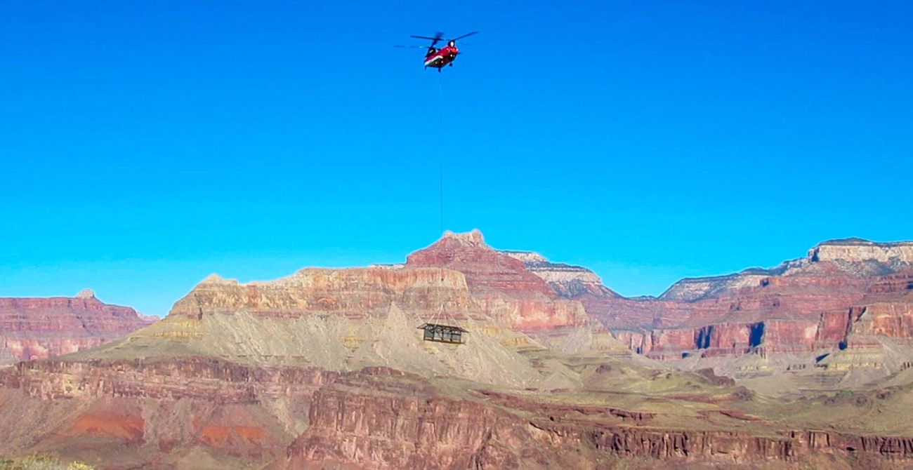 The South Kaibab Trail Tip Off ramada being lowered onto the site. the framework of a 12 x 24 foot shade structure being lowered from a cable by helicopter, with colorful cliffs and peaks in the background.