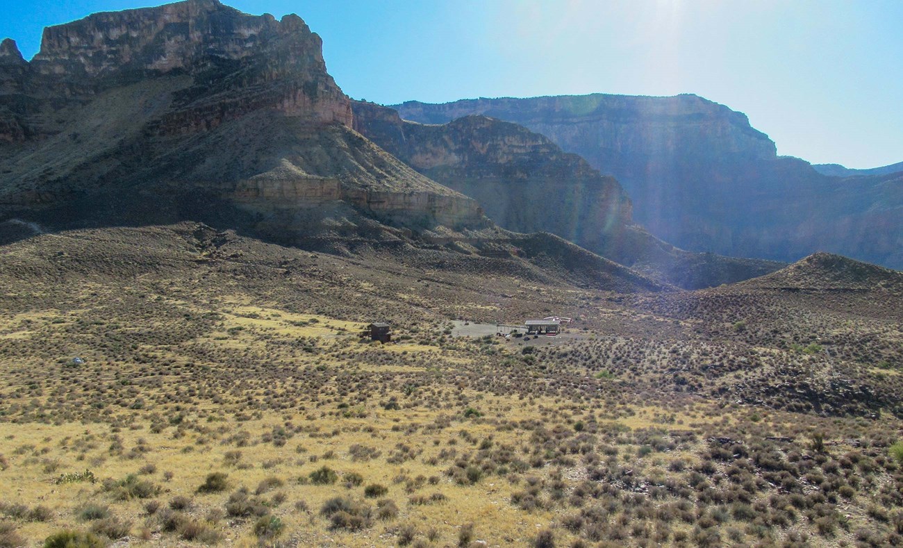 view across a desert plain at a clearing with two small buildings. In the background canyon cliffs rise.