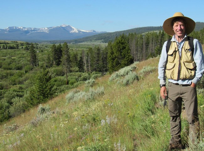Biologist poses by a field of wildfires with scattered trees and snowcapped mountains in the background.