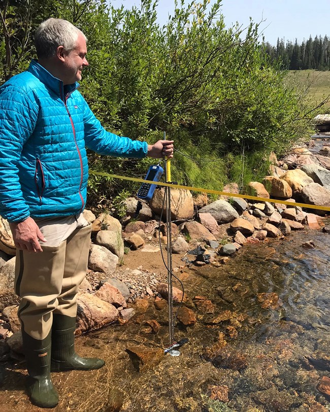 scientist standing on the edge of a creek next to a measuring tape stretching across the creek and holding water quality equipment in the water