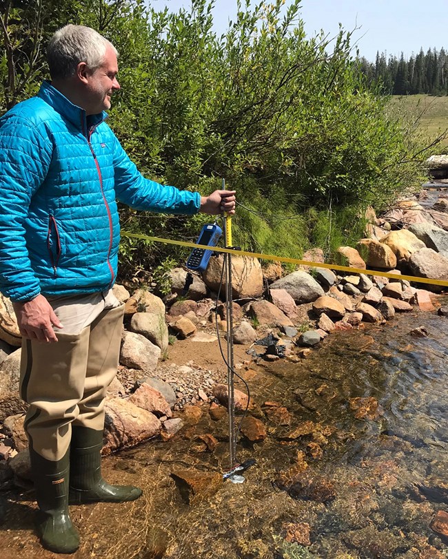 Water quality monitoring in 2018 scientist standing on the edge of a creek next to a measuring tape stretching across the creek and holding water quality equipment in the water