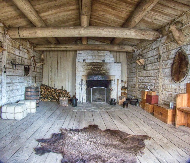 Rectangular room, wooden walls and vaulted ceiling with whitewashed stone fireplace on rear wall and buffalo robe on floor.