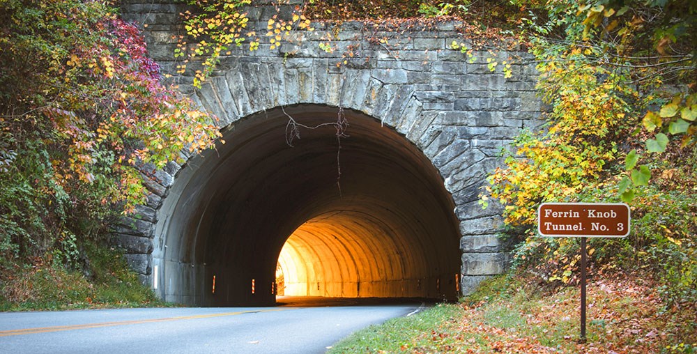 Road running through a stone-lined tunnel. Sunlight glows at the other end of the tunnel.