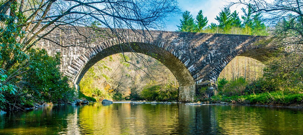 A stone bridge with two arches spans a small river. The bright greens of new, spring leaves are reflected in the water.