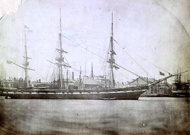 Black and White image: A tall ship without sails site in a harbor.