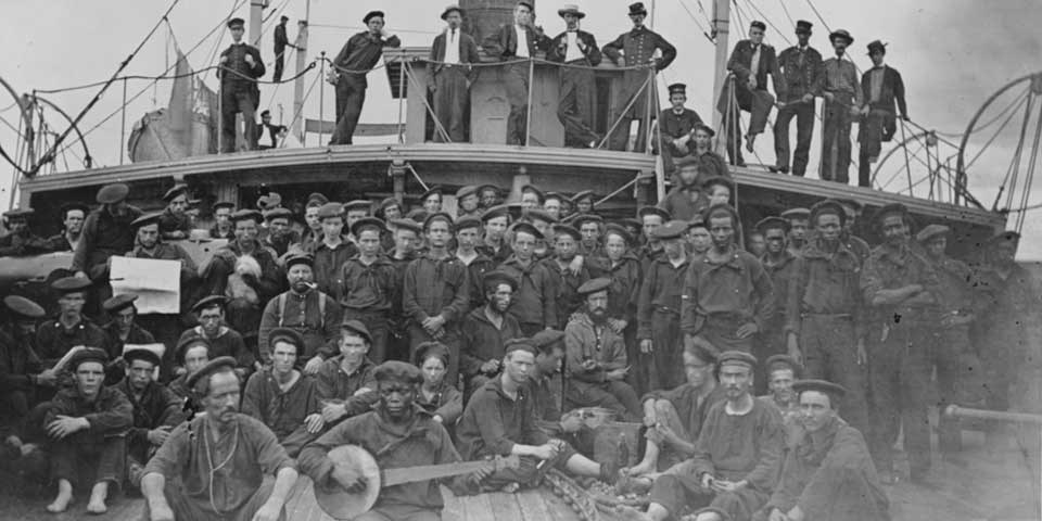 Black and white photograph of the crew aboard USS Hunchback. Some officers stand on a poop deck while most stand or sit on the main deck.