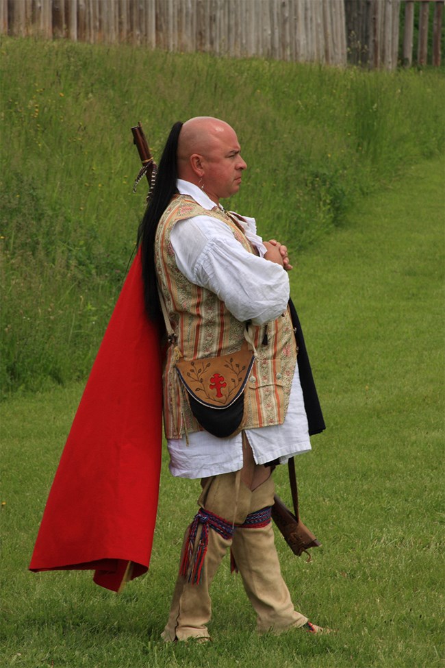 A man in traditional Oneida clothing. A flowing red cape, a yellow-striped vest, feathers adorning his head, and leather leggings.