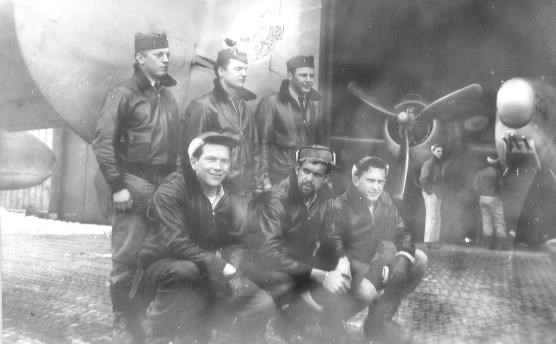 six men standing near a plane, with two more planes and people in hangar behind them.