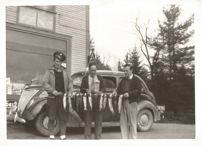 Vincent and his family enjoying Vermont fishing Black and white photo of three men holding a stick with a dozen fish hanging down vertically, in front of an old-fashioned car and building.