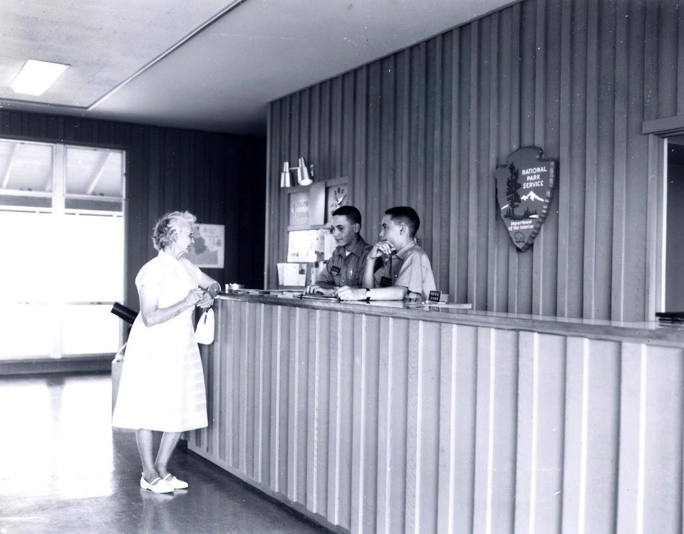 Photo of a front desk with two young men behind it. On the wall behind them is a large arrowhead. A woman stands in front of the desk.