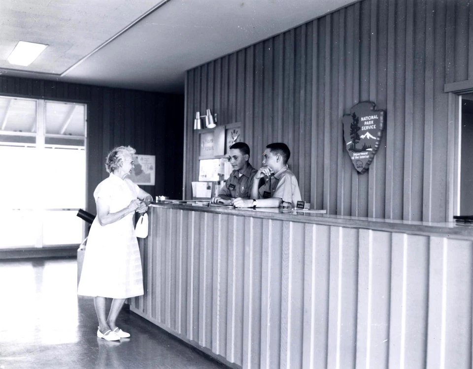 Visitor Center - 1965 Junior Rangers at Visitor Center Photo of a front desk with two young men behind it. On the wall behind them is a large arrowhead. A woman stands in front of the desk.