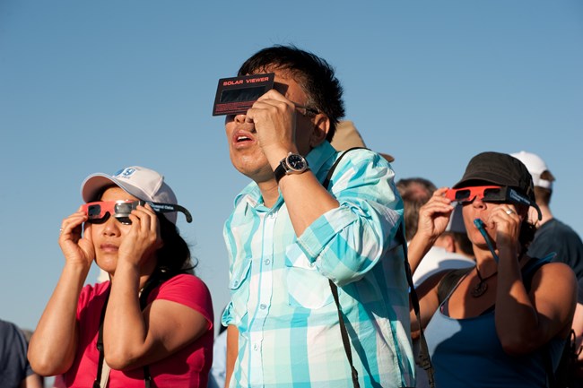 Three visitors viewing an eclipse with solar filters. Two visitors are covering their eyes with eclipse glasses and one visitor is cover their eyes with a hand-held solar viewer.