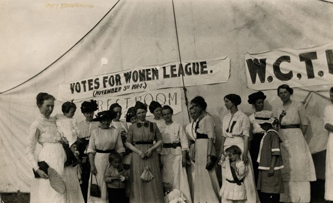 About 16 women and children standing in front of a large tent. Sign reads "Votes for Women League, November 3, 1914"