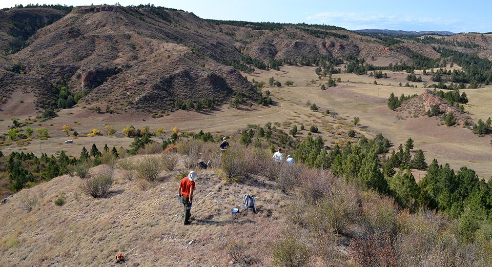 Two people standing by a measuring tape on a hilltop overlooking a valley and more hills, and two people walking down the hill.