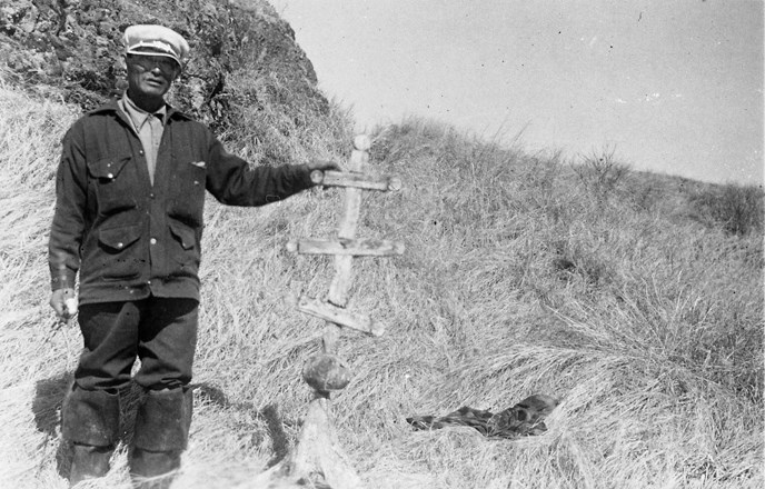 A black and white photograph of a man in a grassy area next to a Russian orthodox style cross A black and white photograph of a man in a grassy area next to a Russian orthodox style cross