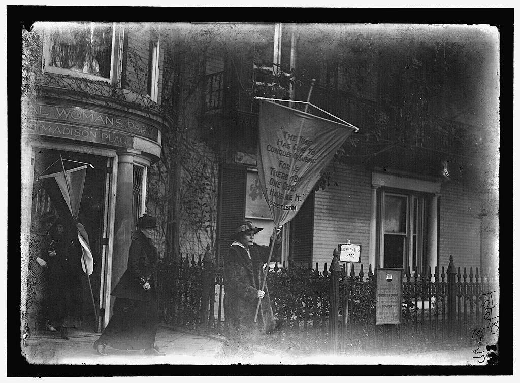 Suffragists walking out of a building. One holds a banner reading, ""The time has come to conquer or submit for there is but one choice--we have made it. -President Wilson"