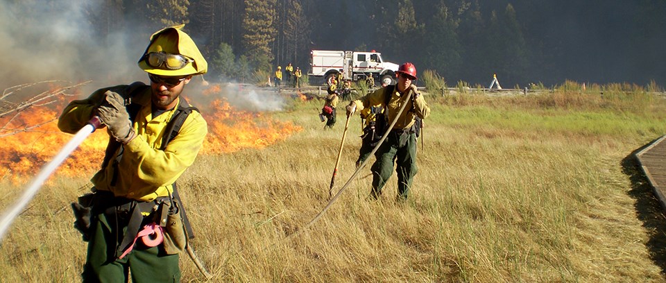A firefighter sprays a hose attached to a wildland fire engine in the distance.