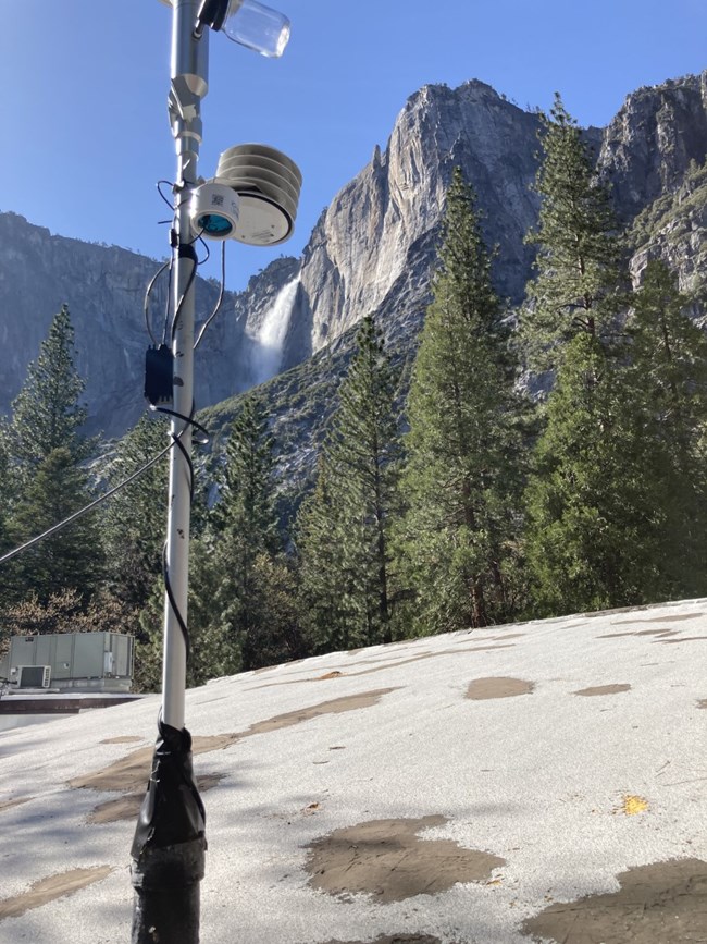 Electrical cords wrap around a silver pole up to an air monitor. In the background is a granite cliff with a waterfall.