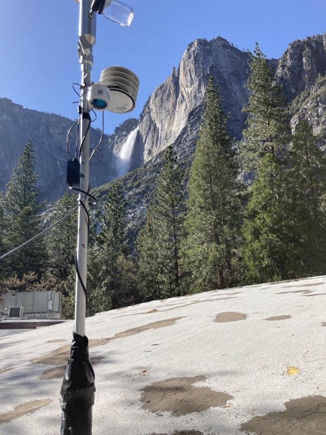 PurpleAir monitor at Yosemite Electrical cords wrap around a silver pole up to an air monitor. In the background is a granite cliff with a waterfall.
