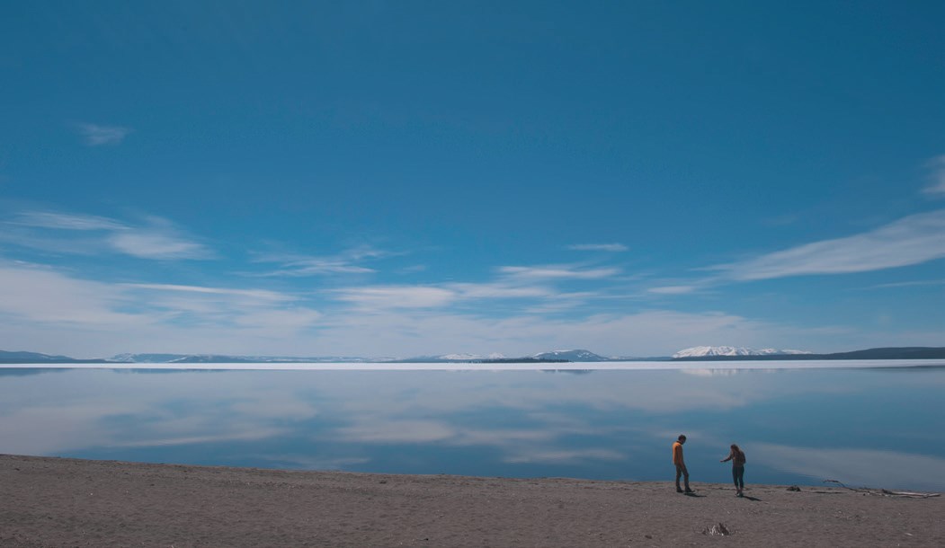 Two people on the beach of a very large lake with snow-capped mountains in the background.
