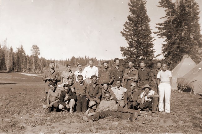 Group of men in a old photo standing in a field with trees in the background.