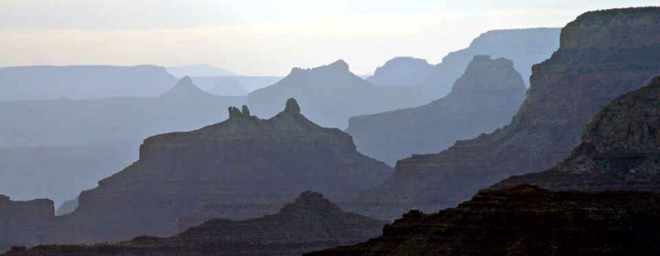 Silhouettes of Grand Canyon rock formations as golden light filters through them. Darker clouds hang overhead.