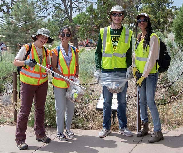 4 college students wearing yellow safety vests are picking up litter.