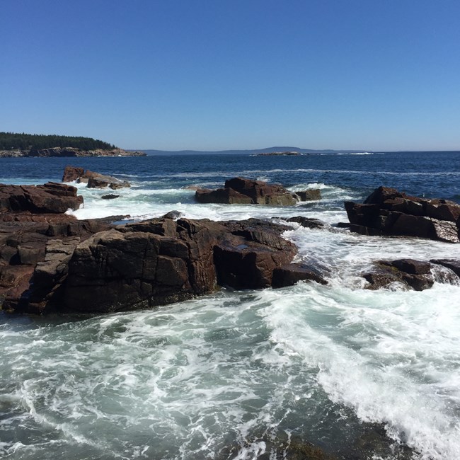Acadia National Park, Maine Under a blue sky ocean waves swell into ragged rocky coast.