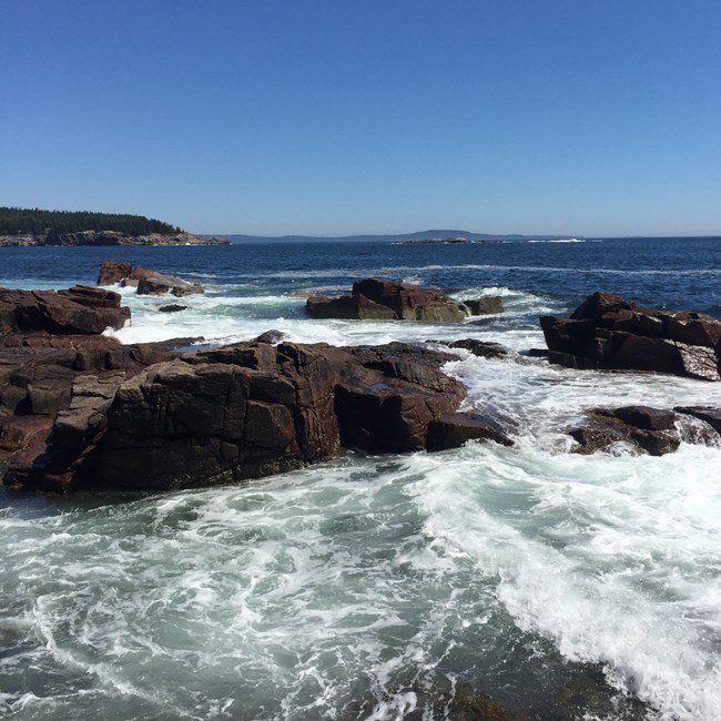 Under a blue sky ocean waves swell into ragged rocky coast.