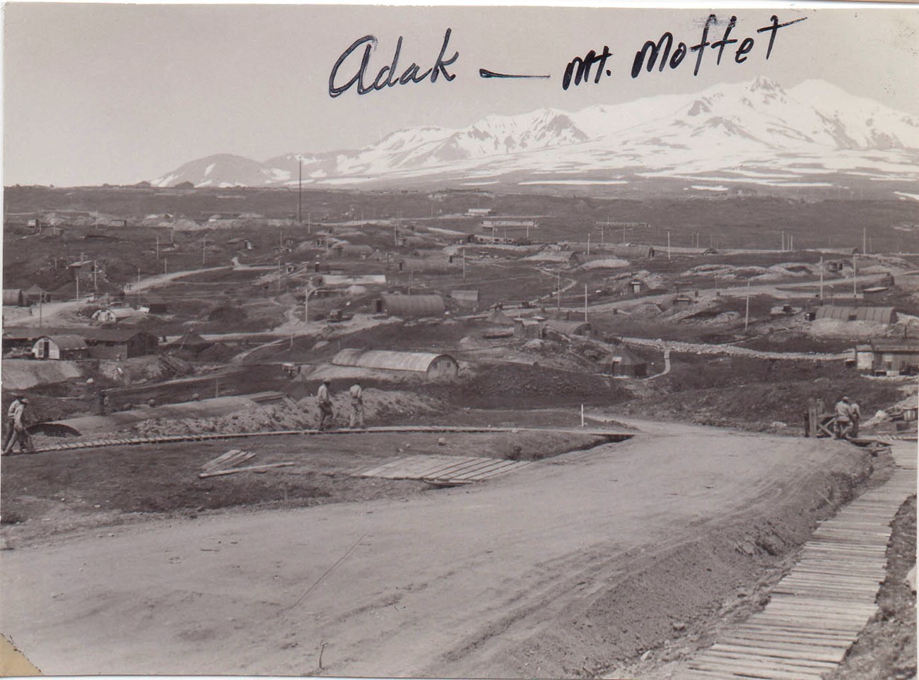 Adak and Mt. Moffet Long dirt ramp with quonset huts and roads in the middle of the image. A large snowy mountain is in the background.