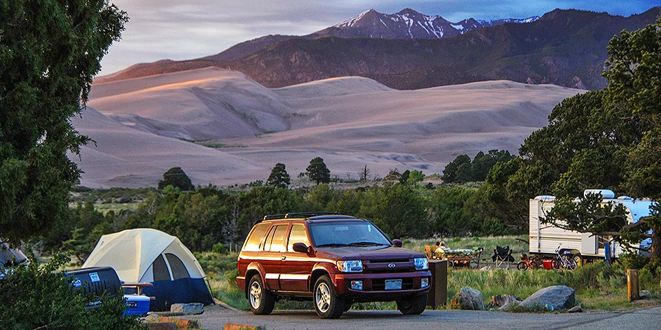 A campsite is seen with tents, cars, and campers, with mountains in the distance