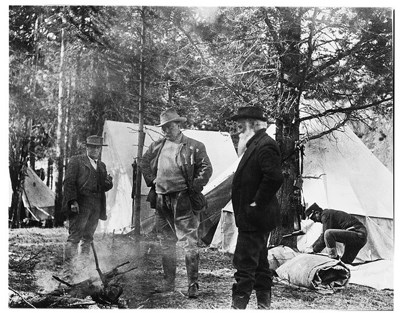 Black and white photo of Teddy Roosevelt standing with other men around a smoldering pile of branches Black and white photo of Teddy Roosevelt standing with other men around a smoldering pile of branches