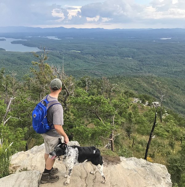 A man and a dog standing on a rock overlooking a green valley. Mountain ridges in the far distance.