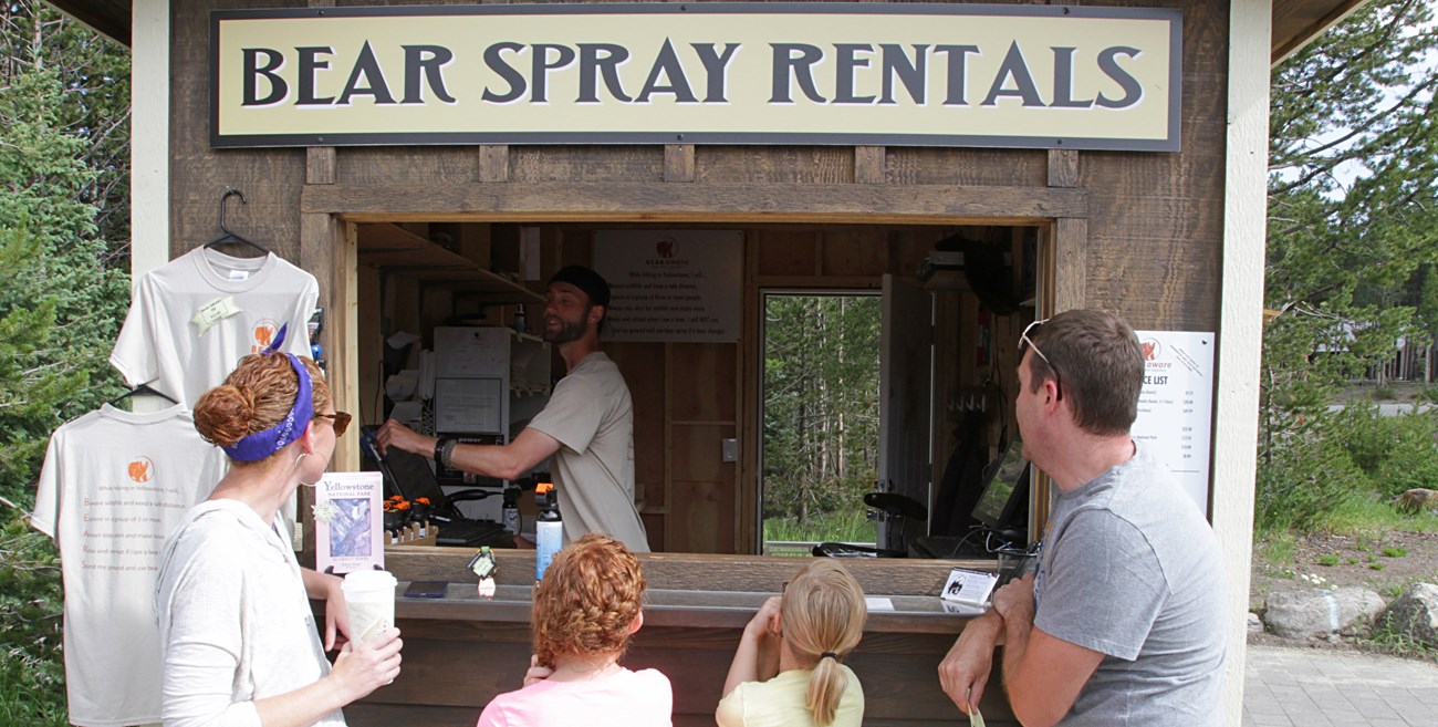 A family of 4 stand at a booth with a sign that reads "Bear Spray Rentals"