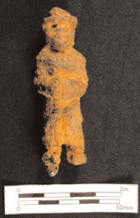 A dirt-covered iron figurine of an African American man, about 3.5 inches tall. The man has his hands held together and wears a hat.